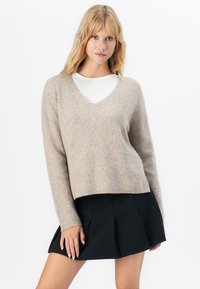 Mujer rubia vistiendo un suéter de pico beige de canalé sobre una blusa blanca y una falda corta plisada negra, posando contra un fondo blanco.