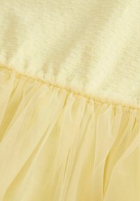 Vêtement jaune en tissu doux et texturé avec un volant en tulle transparent à l'ourlet. Le design est léger et délicat.