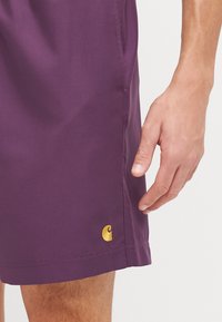 Ljubičaste hlačice od glatkog materijala imaju zlatni logo blizu rubova. Dizajn ima jednostavan kroj s elastičnom gumom u struku.