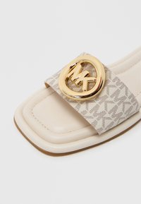 Slip-sandal med en beige mønstret rem med et gyldent rundt logo. Firkantet tå-design med en glat beige sål og syningsdetaljer.