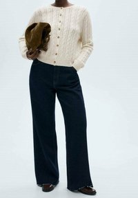 Cárdigan de punto color crema con un patrón texturizado y botones en la parte delantera. Combinado con vaqueros de pierna ancha azul oscuro y mocasines marrones, sosteniendo un sombrero marrón.