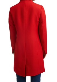 Rode op maat gemaakte jas met een gestructureerde afwerking, een split aan de achterkant, lange mouwen en een scherp gedefinieerde kraag, gecombineerd met een blauwe spijkerbroek.