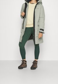Veste imperméable vert clair avec capuche, accents noirs, portée sur un sweat-shirt beige, associée à des leggings vert foncé et des chaussures de randonnée marron.