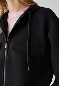 Felpa nera con zip, realizzata in tessuto morbido, con cappuccio, due tasche anteriori con contorno cucito e cordoncini con punte in metallo.