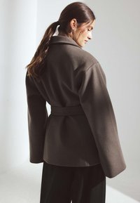 Brūns vilnas maisījuma mētelis ar platu apkakli, jostas daļu un garām piedurknēm. Gluda tekstūra, piegriezums, kas piestāv, un minimālisma dizains. Muguras skats.