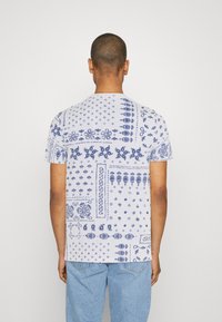 Valkoinen lyhythihainen T-paita, jossa on monimutkaisia sinisiä kuvioita, mukaan lukien kukka- ja geometrisia aiheita, pyöreällä kaula-aukolla.