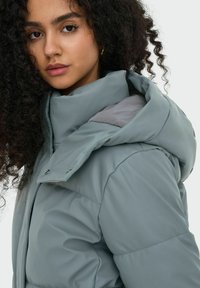 Chaqueta acolchada de color azul verdoso con un alto cuello, que cuenta con una capucha desmontable y forro gris. Textura suave y broches de presión plateados.