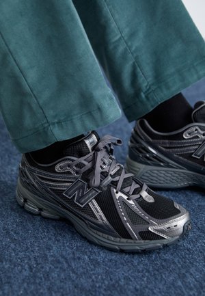 Personne portant des baskets New Balance noires et argentées avec des lacets gris, des chaussettes noires et un pantalon bleu sarcelle, debout sur un sol moquetté bleu.