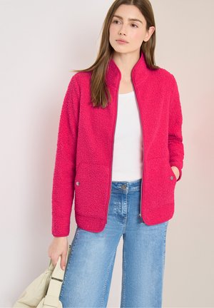 Frau trägt eine leuchtend pinke Fleecejacke, weißes Oberteil und blaue Jeans, hält eine beige Tasche vor einem schlichten hellen Hintergrund.