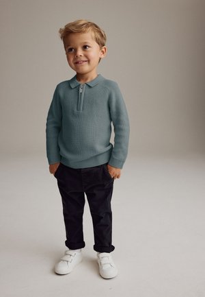 Nuori poika seisoo käsivarret taskuissa, päällään merensininen vetoketjullinen neulepaita, mustat housut ja valkoiset lenkkarit, yksinkertaisen taustan edessä.