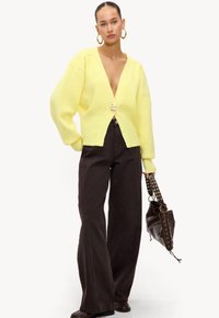 Geel gebreid cardigan met een diepe V-hals, oversized mouwen en een grote gouden knop. Gecombineerd met donkerbruin brede broek en een bruine tas.