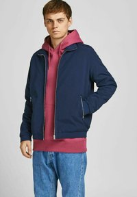 Marine bomberjack met ritssluiting, gecombineerd met een roze hoodie. Gedragen met lichtblauwe jeans, met een soepele, matte textuur.