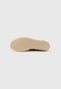 Semelle de chaussure en caoutchouc beige texturé, dotée de motifs circulaires et du nom de la marque gravé. Le bord supérieur se courbe doucement, passant au rouge.