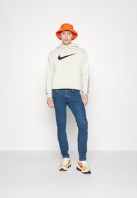Jovem de chapéu laranja em forma de balde, com um hoodie branco da Nike, jeans azuis e ténis laranja e brancos, em pé contra um fundo branco.