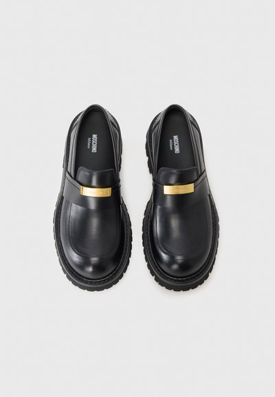 Svarta läderloafers med en slät yta, prydda med ett guldtonat accentband över framsidan och en texturerad gummisula.