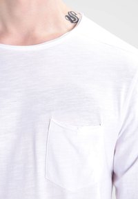 Weißes T-Shirt mit kurzen Ärmeln aus leichtem Stoff, mit einer Brusttasche und rundem Ausschnitt. Glatte Textur, minimales Design.
