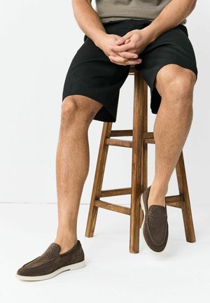 Mies, jolla on mustat shortsit ja ruskeat liukukengät, istuu puisella jakkaralla kädet sylissä.