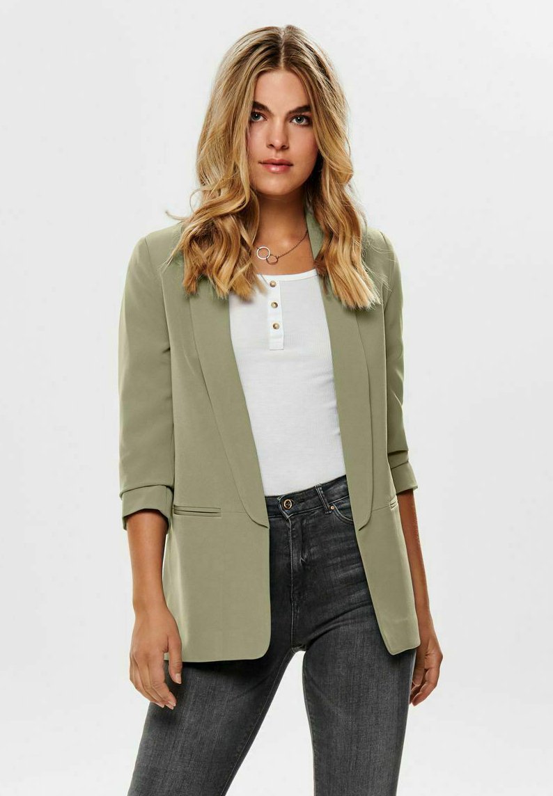 Blazer vert clair avec revers crantés, deux poches avant et manches retroussées, associé à un haut blanc à boutons et un jean foncé.