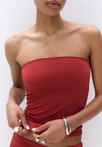 Rød stropløs top lavet af glat stof, med en tætsiddende design. Person bærer sølvringene og armbånd, med en simpel baggrund.