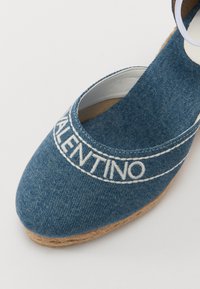 Denimové espadrilky s kulatou špičkou, zdobené bílým vyšívaným textem a středovou podrážkou obalenou jutou. Světle modrá barva a hladká textura.