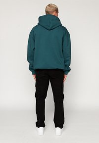 Teal Hoodie mit einer geräumigen Kapuze und gerippten Bündchen, kombiniert mit schwarzen Cargo-Hosen und weißen Sneakers. Der Stoff wirkt weich und texturiert.
