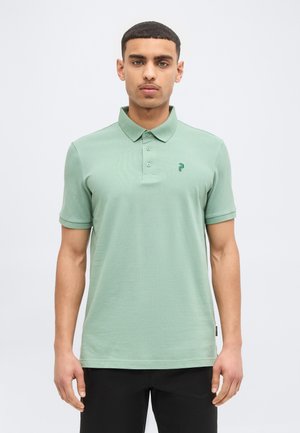 Uomo con capelli corti che indossa una polo a maniche corte verde chiaro e pantaloni neri, in piedi contro uno sfondo bianco semplice.