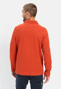 Langærmet polo skjorte i lys orange bomuld. Har en krave, sidesplitter og et enkelt design. Bæres med mørke bukser.