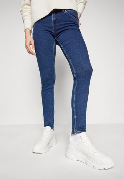 Vaqueros ajustados de mezclilla azul con una textura suave, decorados con costuras naranjas, combinados con zapatillas deportivas gruesas blancas.
