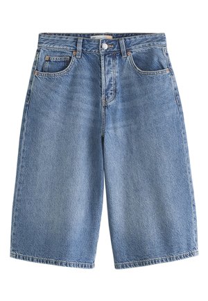 Frau trägt hochtaillierte, locker sitzende knielange Jeansshorts und ein rotes Crop-Top, steht nahe weißen Treppen mit Topfpflanzen.