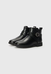 Bottines en cuir noir avec un bout arrondi, détail de sangle avec une boucle circulaire, et une semelle plate et basse. La texture est lisse et brillante.