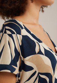 Bluse mit floralem Muster in Marine und Beige, mit V-Ausschnitt, kurzen Ärmeln und weichem, strukturiertem Stoff. Das Design zeigt große Blätter.