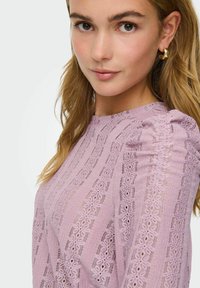 Ung kvinne med lysebrunt hår iført gull hoops øredobber og en rosa blondertop med puffermer, vendt mot kameraet.