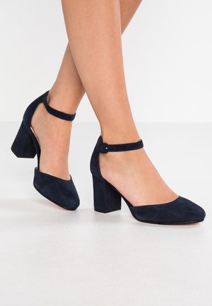 Woman wearing navy blue suede block heel shoes with ankle straps standing on a white floor.