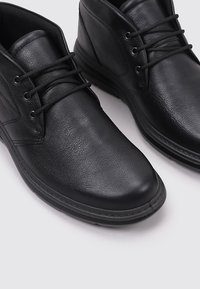 Las botas de tobillo de cuero negro tienen una textura suave, punta redonda y un sistema de cordones con seis ojales. Suela de goma negra sólida para mayor estabilidad.