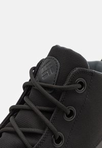 Svart kanvas sneakers med teksturert overdel, runde svarte øyetter og flate lisser. Tungen har en subtil preget logo detalj.