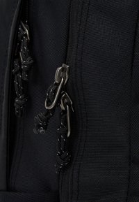 Černý látkový batoh s dvojitými kovovými zipy a černými šňůrkami, texturovaný povrch, viditelné švy a zesílené švy.