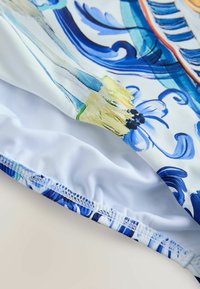 Maudymosi kostiumėlis su daugiacolore gėlių raštu mėlynos ir geltonos spalvos, su lygiu balto audinio vidumi ir elastiniu siuvimu kraštuose.