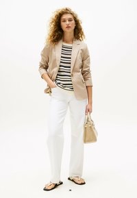 Blazer beige sopra una maglietta a righe, abbinato a pantaloni bianchi. Gli accessori includono una piccola borsa beige e ciabatte nere su uno sfondo chiaro.