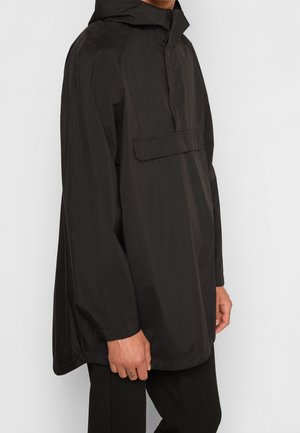 Mann trägt einen lockeren schwarzen Kapuzenpullover mit vorderer Taschenklappe und steht vor einem weißen einfarbigen Hintergrund.