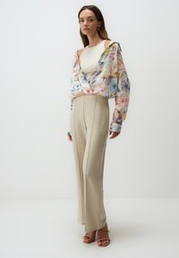 Blusa floreale fluida e trasparente con colori pastello, abbinata a pantaloni beige a vita alta e sandali con tacco di colore chiaro.