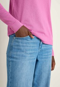 Rosa, taillierter Pullover mit langen Ärmeln, kombiniert mit hellblauen Jeans. Der Pullover hat eine glatte Textur; die Jeans sind leicht verwaschen und haben Taschen.
