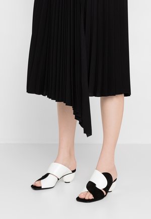 Pieds portant des sandales à talons ouvertes noires et blanches, associées à une jupe midi plissée noire avec un ourlet asymétrique.