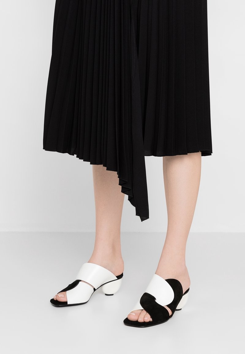 Pieds portant des sandales à talons ouvertes noires et blanches, associées à une jupe midi plissée noire avec un ourlet asymétrique.