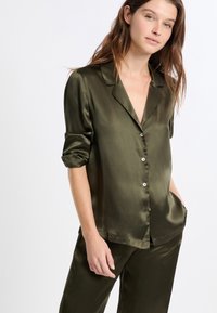 Conjunto de pijama de satén verde oliva que incluye una blusa abotonada con cuello, mangas remangadas y pantalones a juego con bolsillos laterales. Textura suave.