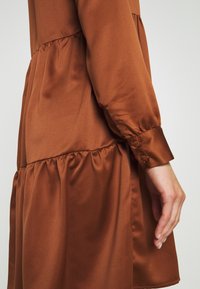 Brun satin klänning med ett lager- design, långa ärmar med knäppta mudd, och samlat detaljer vid midjan för extra textur.