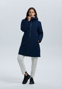 Marineblauwe lange jas met een capuchon, ritssluiting, zijzakken en een gladde textuur. Gepaard met witte broeken en zwarte schoenen.