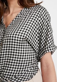 En närbild av en kvinna i en svart och vit rutad skjorta som visar V-ringning och uppvikt ärmar, med en ljus beige tyg nedanför.