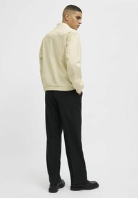 Veste beige clair avec un col, un ourlet élastiqué et des manches longues, associée à un pantalon noir et des chaussures noires. Design simple et fonctionnel.