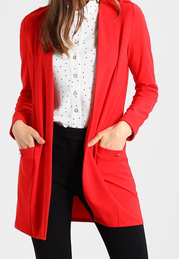 Veste longue rouge en tissu lisse, dotée de deux poches avant et d'un design ouvert. Portée sur une chemise blanche à boutons avec pois.