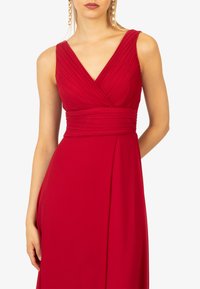 Abito da sera rosso con scollatura a V e corpo plissettato, caratterizzato da una vita aderente e una gonna fluente. Tessuto liscio senza motivi visibili.